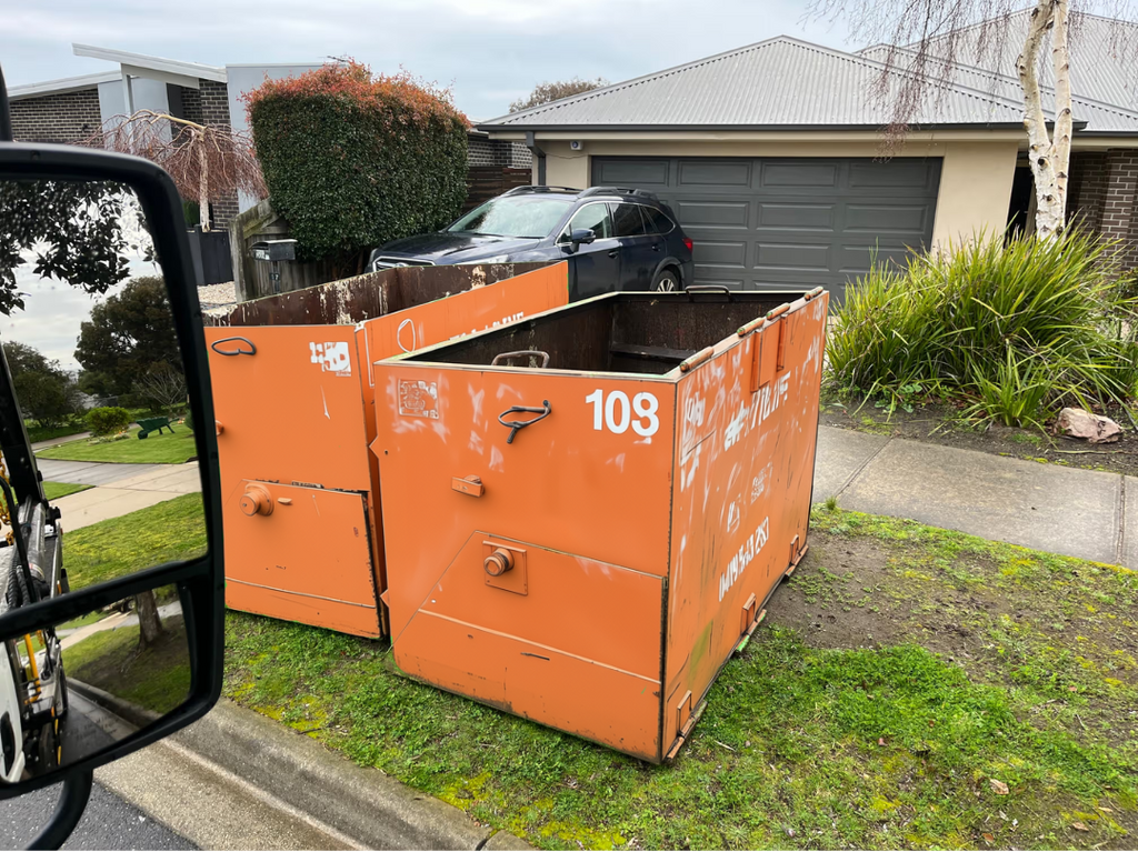 Two orange metal dumpsters with white printed numbers and letters, placed on a grassy area next to a residential street with a garage and house in the background.
