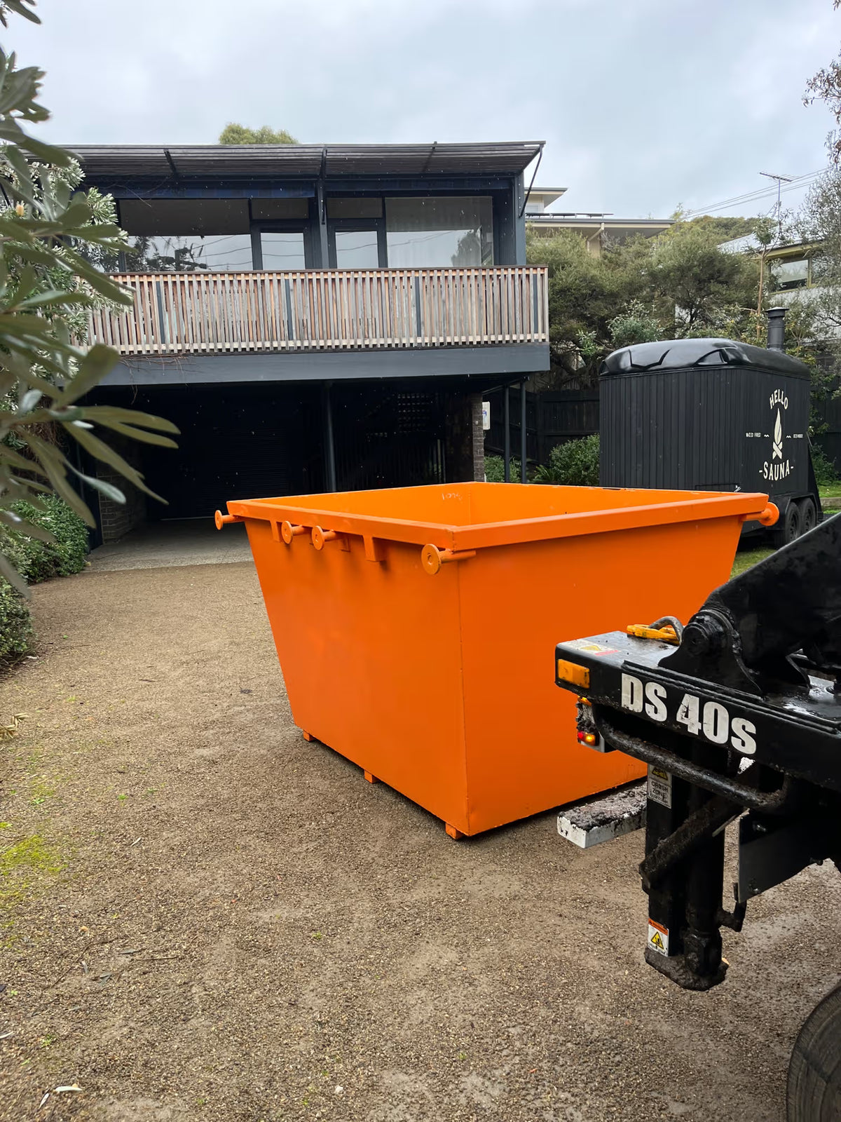 An orange metal dumpster container parked outdoors on a gravel driveway in front of a modern house, with part of a black DS 40S truck visible in the foreground.
