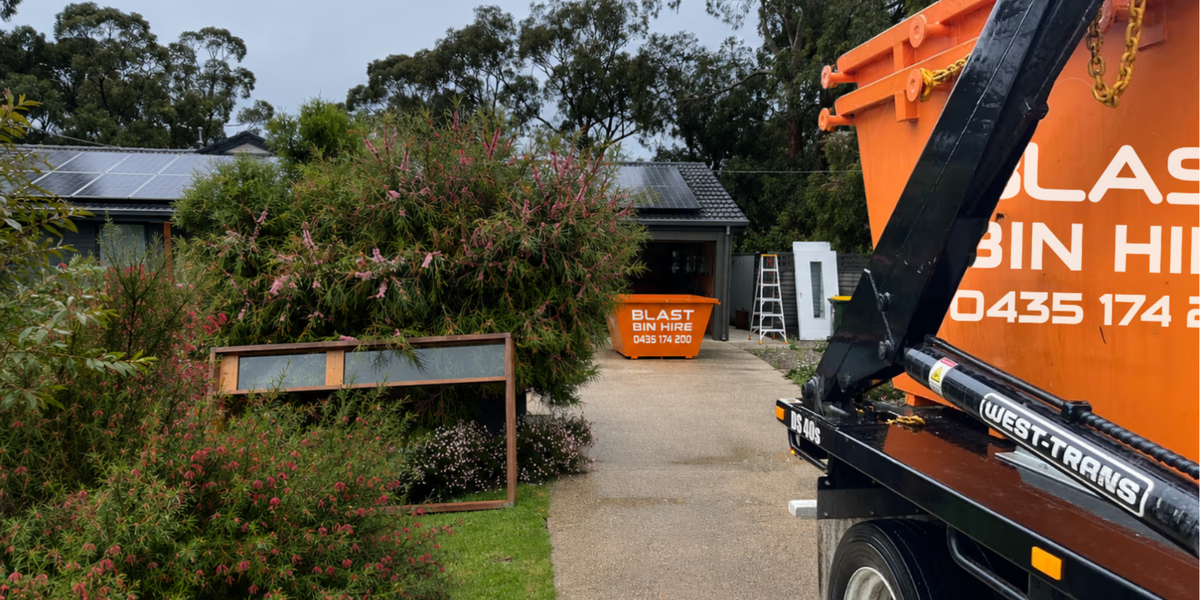 An orange and black Blast Bin Hire dump truck is parked on a driveway in front of a house, with a matching orange dumpster visible in the yard. The scene is outdoors with trees, shrubs, and a house in the background.