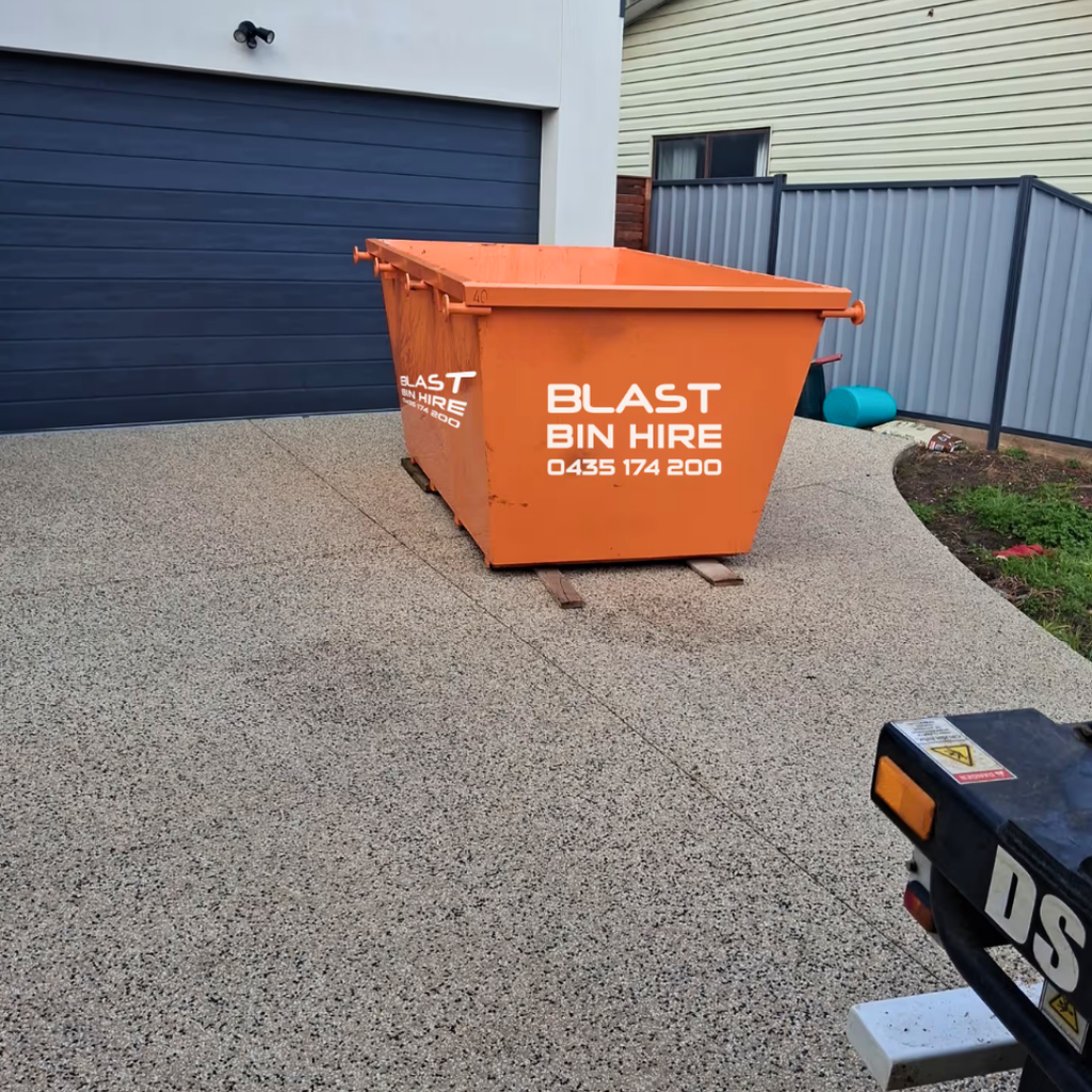 An orange plastic skip bin with 'BLAST BIN HIRE' printed in white on the side, positioned on a concrete driveway in front of a dark blue garage door.