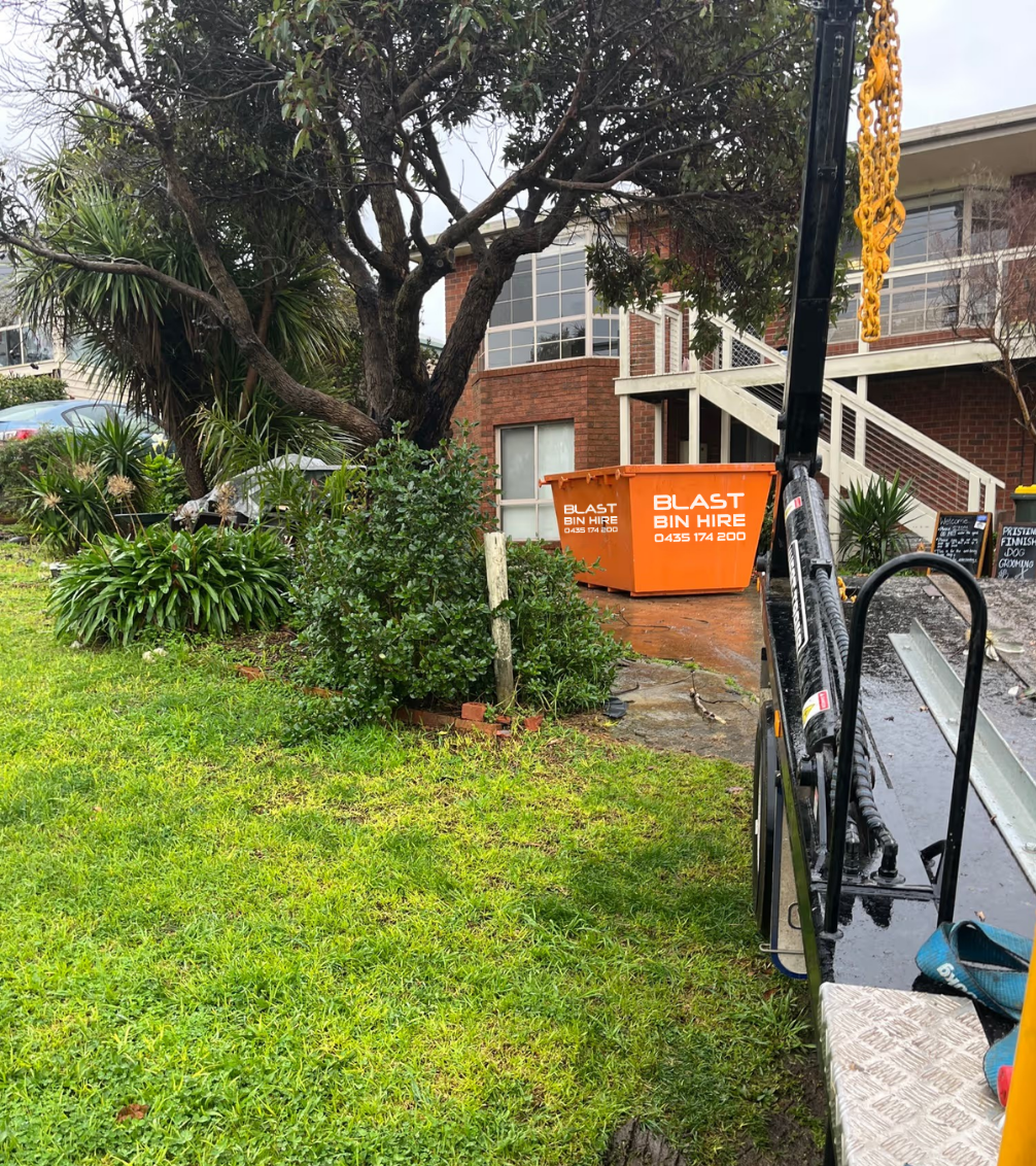 A large orange Blast Bin Hire dumpster sits on a wet paved area next to a grassy lawn and a brick residential building, viewed from the side of a vehicle.