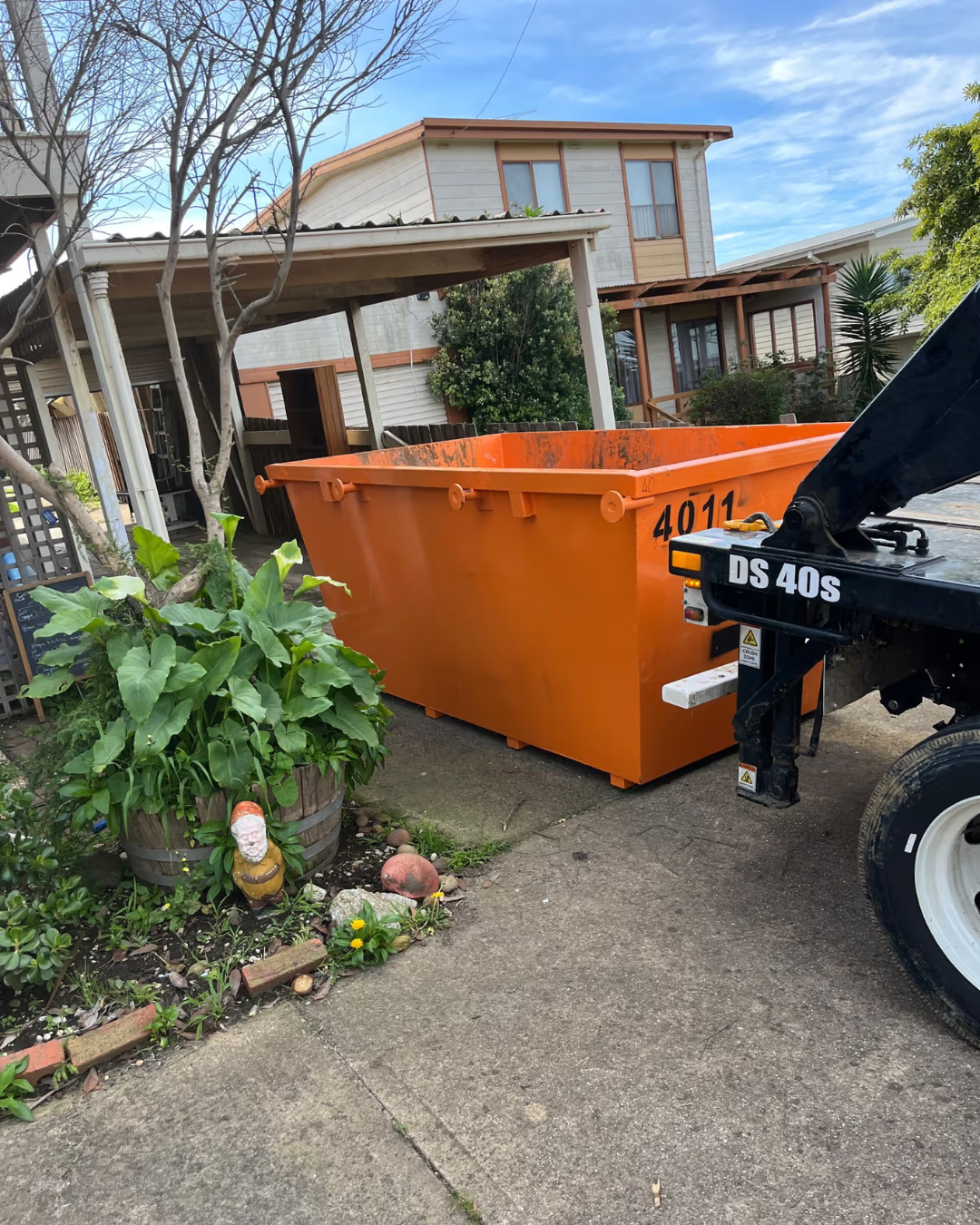 An orange metal dumpster container with the number 4011 on the side, parked outdoors on a concrete driveway next to a black vehicle on a residential street.