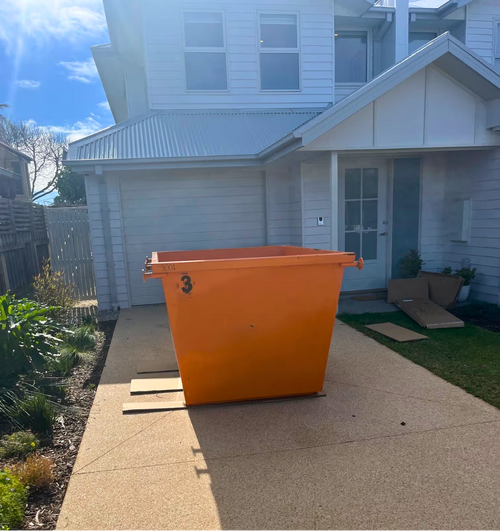 An orange industrial dumpster container placed on a concrete driveway in front of a white house.