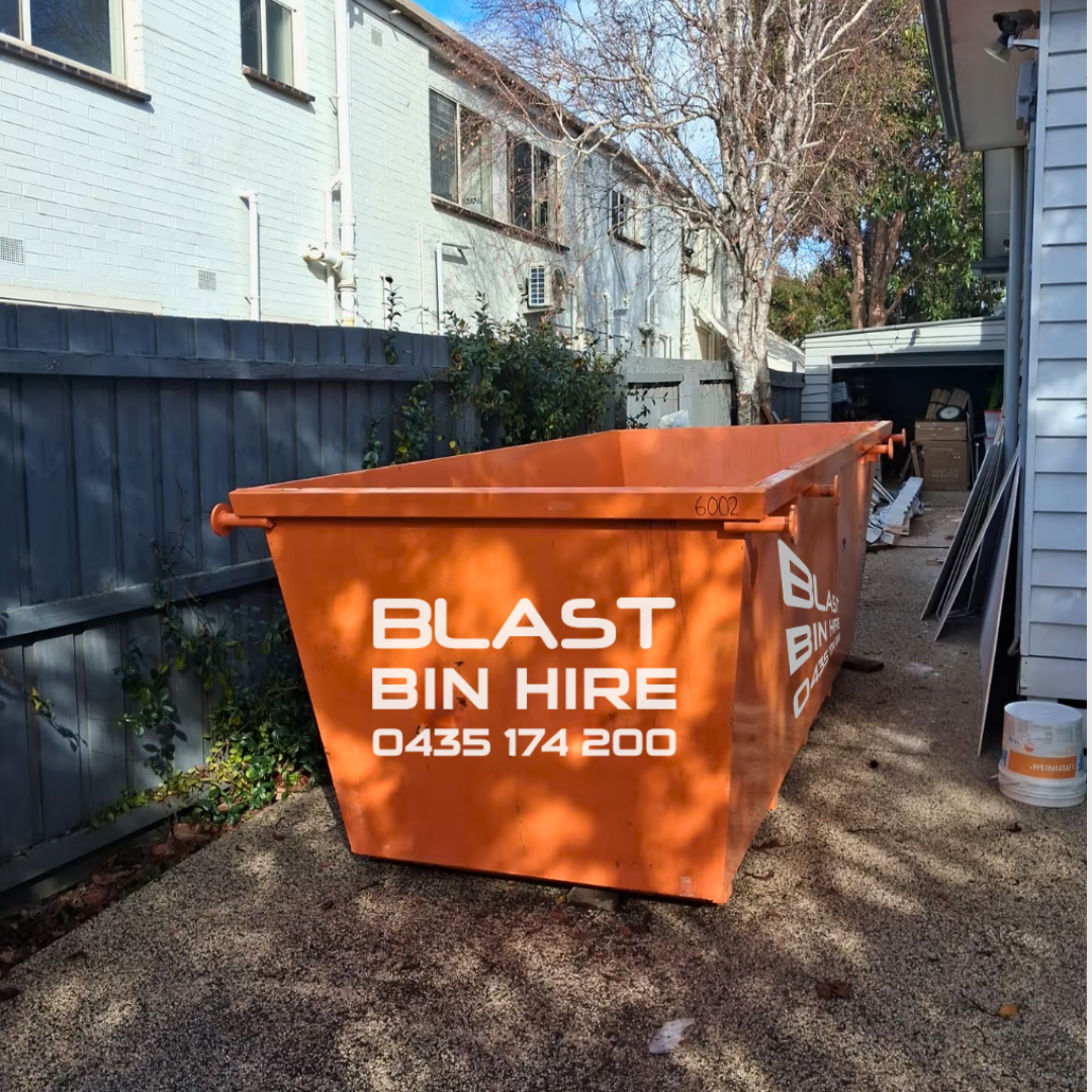 An orange metal skip bin with white text reading 'BLAST BIN HIRE' and a phone number, parked outdoors on gravel between houses.