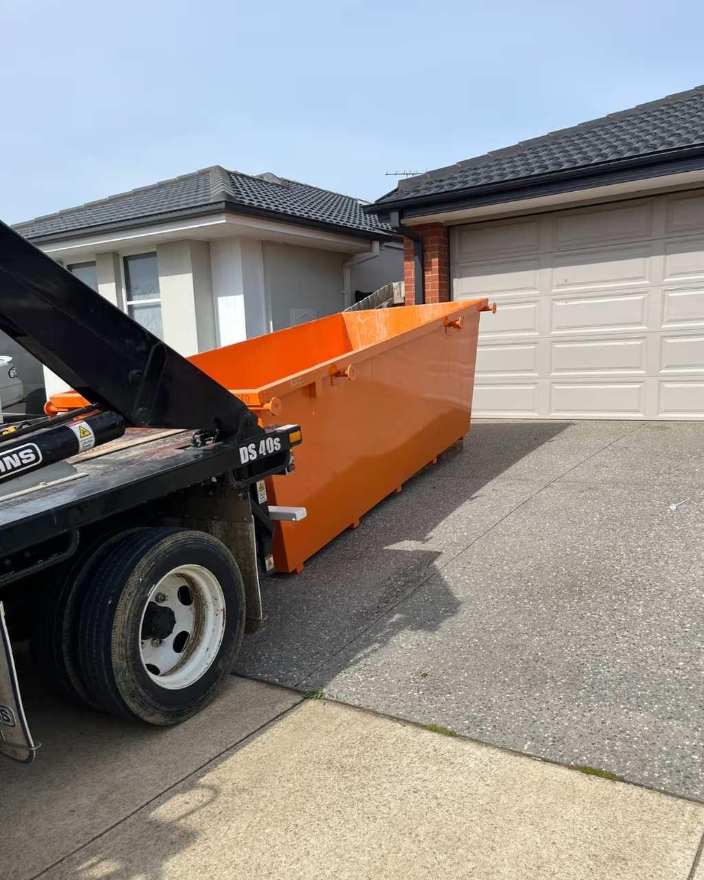 An orange metal dumpster container parked on the side of a black flatbed truck on a residential driveway, with houses and garage doors in the background.