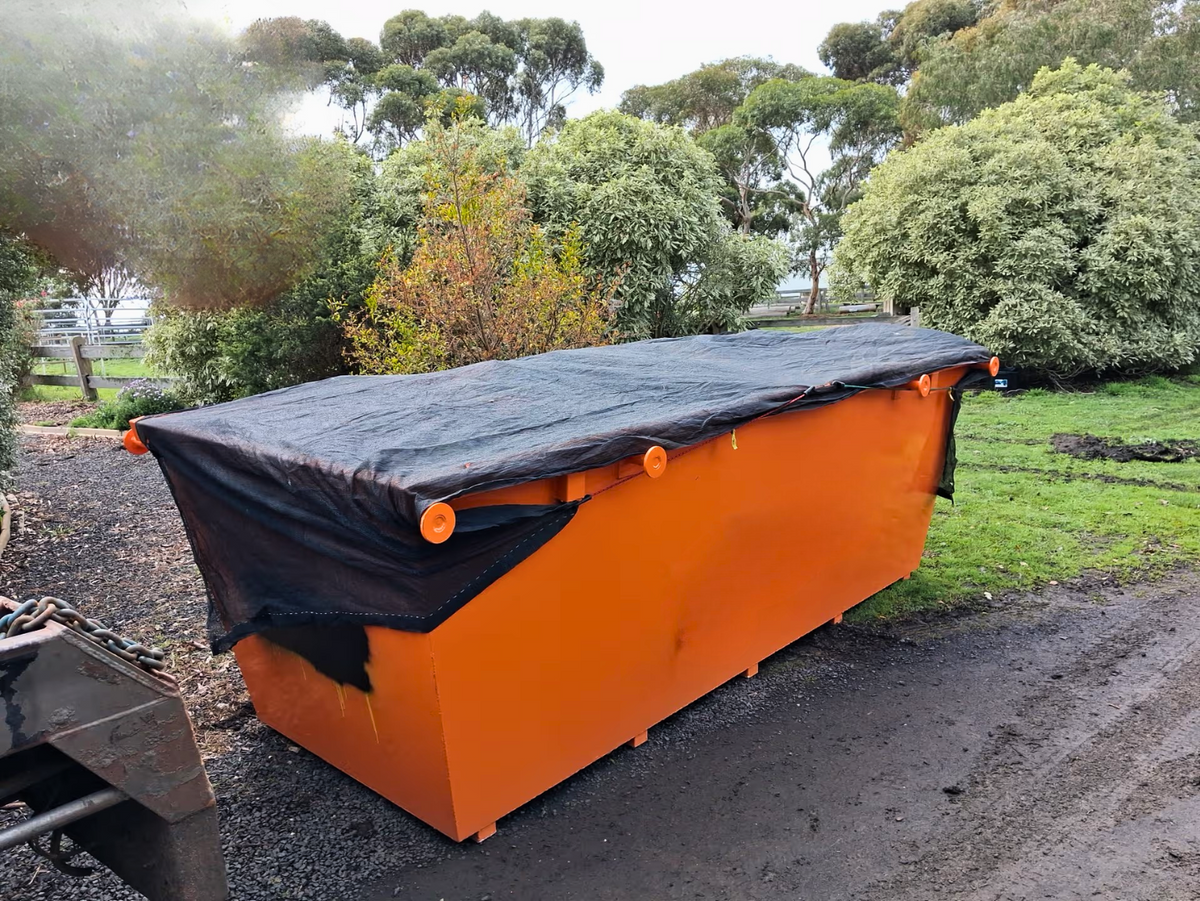 An orange rectangular container or dumpster covered with a black tarp, photographed outdoors on a gravel path with trees and grass in the background.
