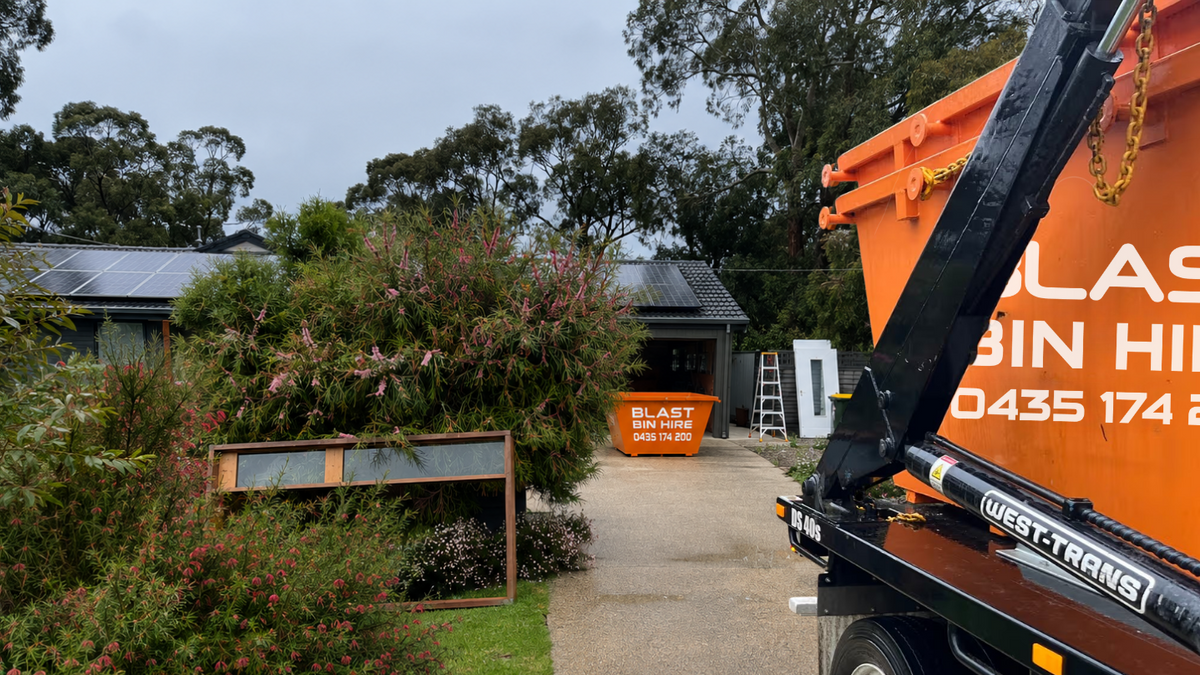 An orange dumpster on a truck parked outdoors beside a house driveway, with another dumpster and garden beds in the background.