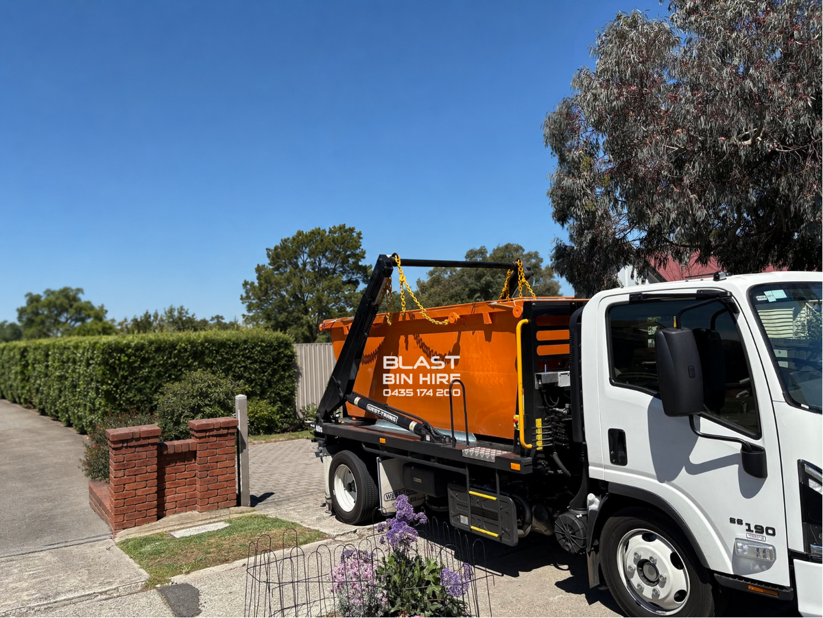 A white cab truck with an orange waste bin mounted on a rear platform, parked outdoors on a driveway beside a hedge and trees under a clear blue sky. The bin has the text 'BLAST BIN HIRE' printed on it.