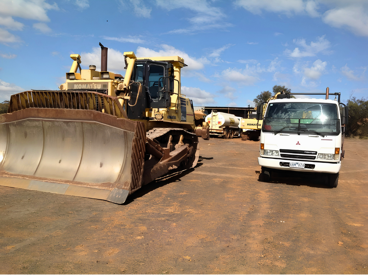 A white Mitsubishi utility truck parked on a dirt lot next to a large yellow CAT bulldozer under a blue sky with clouds.