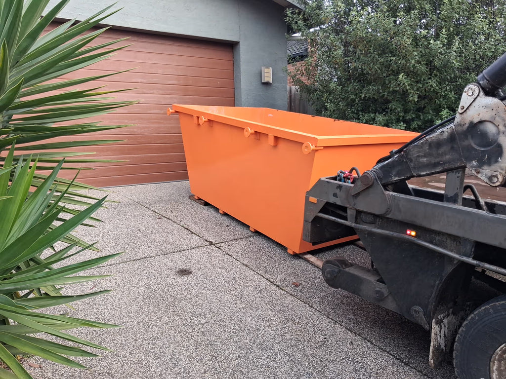 An orange metal dumpster parked on a concrete driveway in front of a brown garage door, with part of a black truck loader visible on the right and green plants in the foreground.