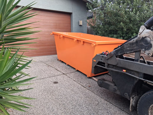 An orange metal dumpster parked on a concrete driveway in front of a brown garage door, with part of a black truck loader visible on the right and green plants in the foreground.